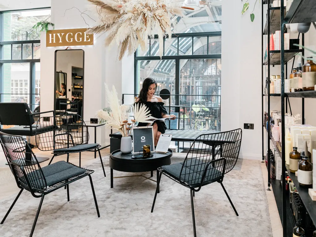 A woman sits in a beauty salon with a sign saying 'Hygge' in the background